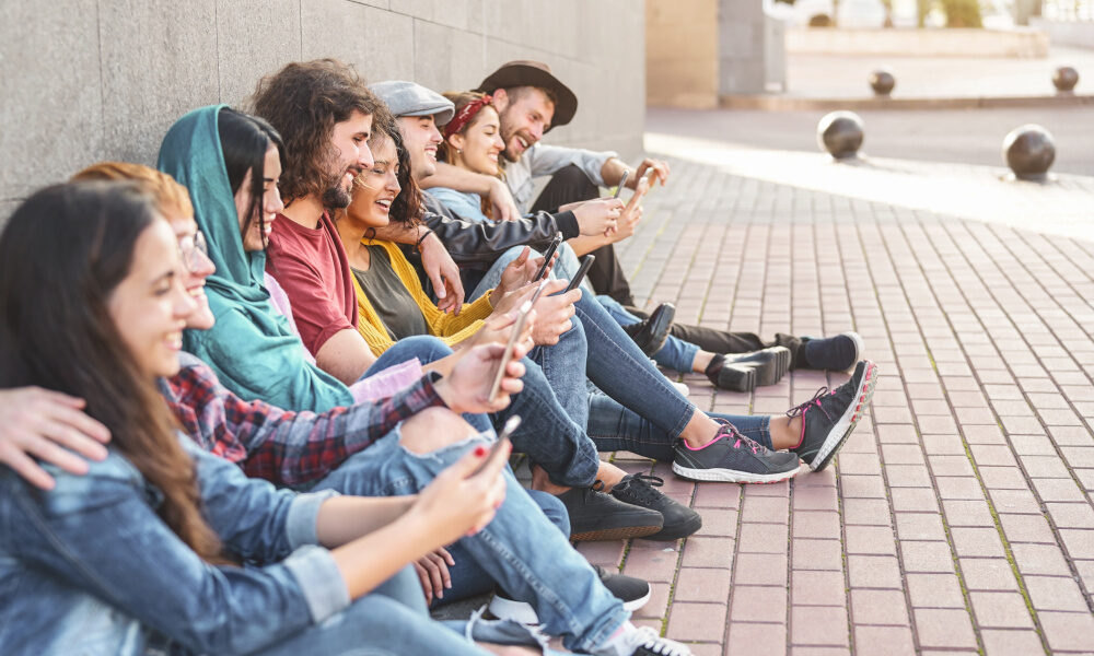 Gruppe junger Erwachsener sitzt entspannt draußen auf dem Boden, eng beieinander, und schaut auf ihre Smartphones.