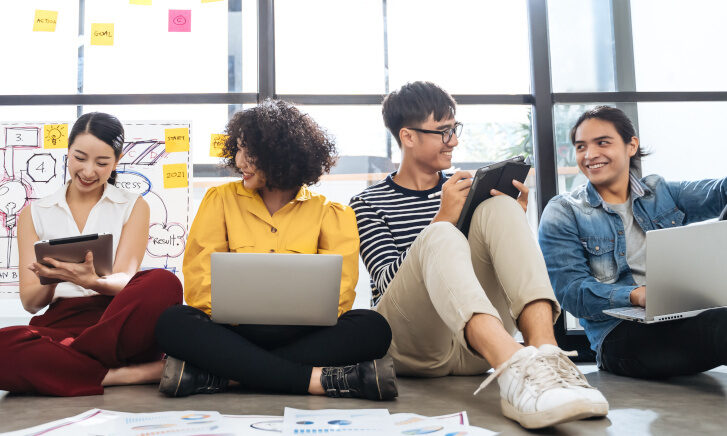 Vier junge Menschen sitzen lachend auf dem Boden mit Laptops und Tablets, diskutieren gemeinsam in lockerer Arbeitsatmosphäre.