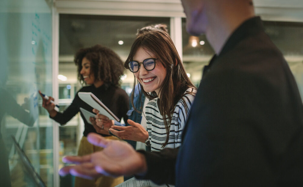 Eine Gruppe junger Menschen arbeitet zusammen an einem Whiteboard, lachend und mit Notizblöcken in der Hand.