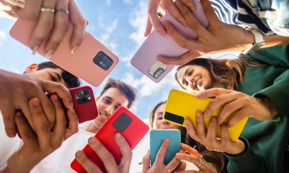 Eine Gruppe junger Menschen hält ihre bunten Smartphones in die Mitte und schaut lachend nach unten.