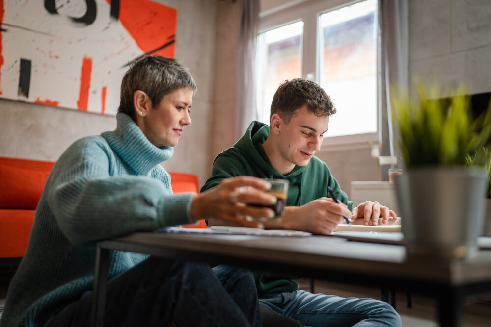 Eine Frau unterstützt einen Jugendlichen beim Lernen, während sie gemeinsam an einem Tisch sitzen.