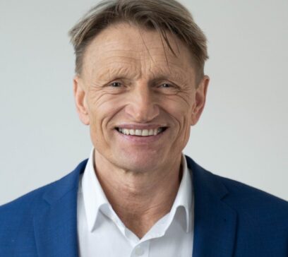 Portrait eines ältereren Mannes mit kurzem Haar, der in die Kamera lächelt. Er trägt ein weißes Hemd und ein blaues Jackett.