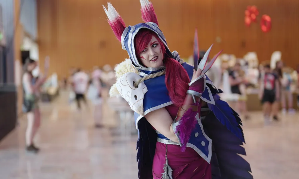 Person in auffälligem Cosplay-Kostüm mit roten Haaren, blauen und weißen Verzierungen sowie großen Ohren posiert auf einer Convention. Im Hintergrund sind verschwommen andere Besucher:innen zu sehen.