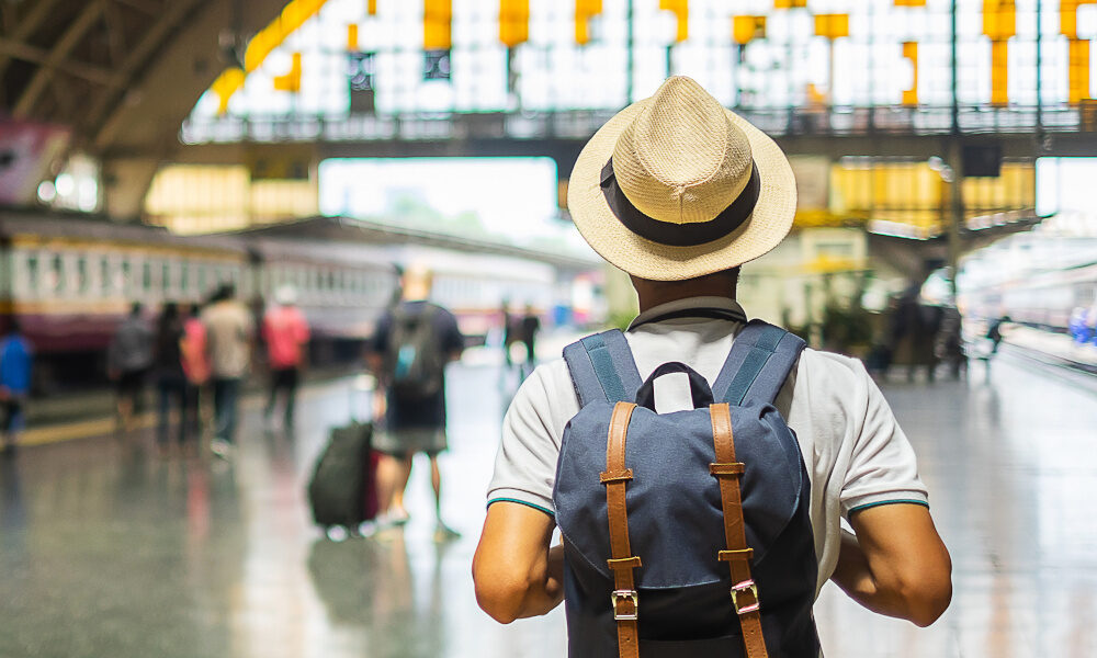 Rückansicht eines jungen Reisenden mit Strohhut und Rucksack, der in einer Bahnhofshalle vor den Gleisen steht.
