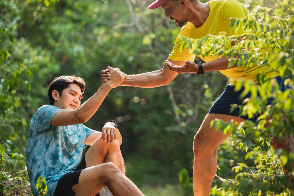 Zwei Männer in Sportkleidung befinden sich im Wald; einer sitzt am Boden und der andere hilft ihm mit einem festen Händedruck hoch.