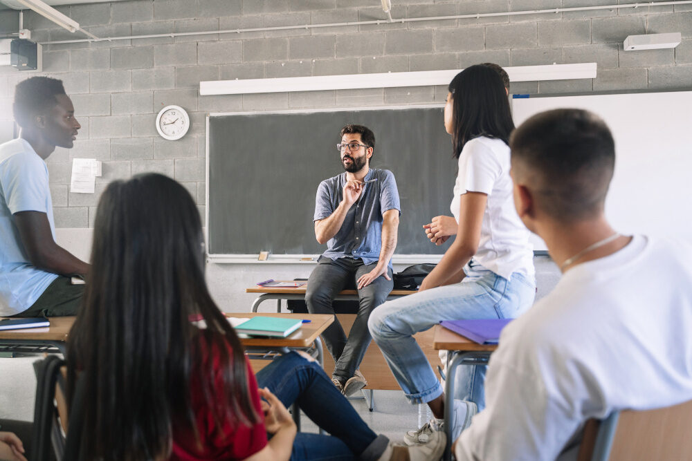 Eine Gruppe von Schüler:innen sitzt in einem Klassenzimmer um einen Lehrer herum, der auf einem Tisch sitzt und gestikuliert. Die Jugendlichen hören aufmerksam zu und diskutieren gemeinsam.
