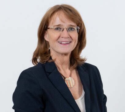 Das Portrait einer Frau mit hellbraunen Haaren und Brille, die einen schwarzen Blazer trägt und in die Kamera lächelt.