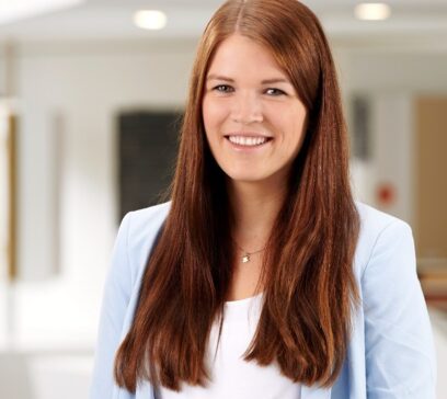 Eine junge Frau in einem weißen Hemd mit einer hellblauen Jacke und braunen Haaren lächelt in die Kamera. Dahinter ist unscharf eine Büroumgebung zu sehen.