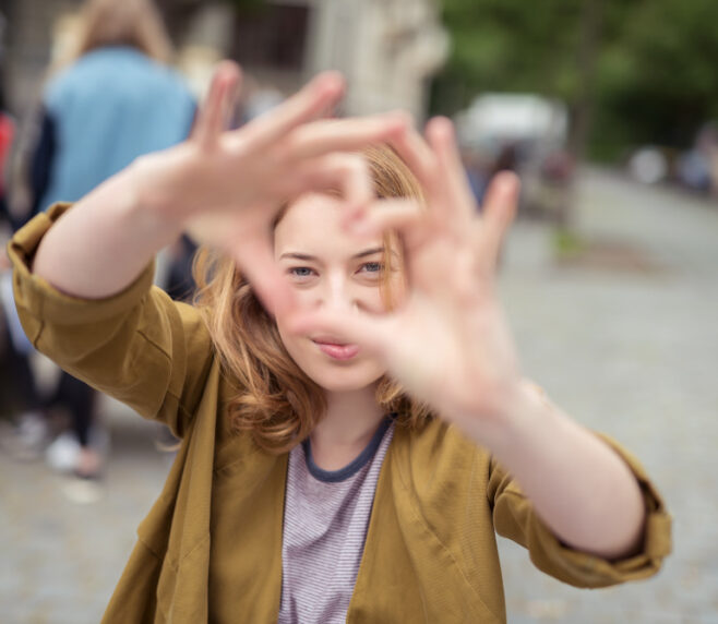 Junge Frau mit blonden Haaren und einem gelben Hemd hält die Hände hoch, um mit ihren Fingern ein Herz zu formen. Im Hintergrund ist verschwommen eine Straße mit Pflasterstein mit Bäumen und ein paar anderen Menschen zu sehen.