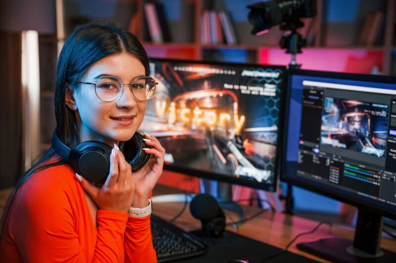 Junge Frau mit Brille und Kopfhörern um den Hals sitzt vor zwei Monitoren in einem Gaming-Setup. Auf einem Bildschirm ist groß das Wort „Victory“ zu sehen. Im Vordergrund liegen ein Mikrofon und Technik-Equipment.