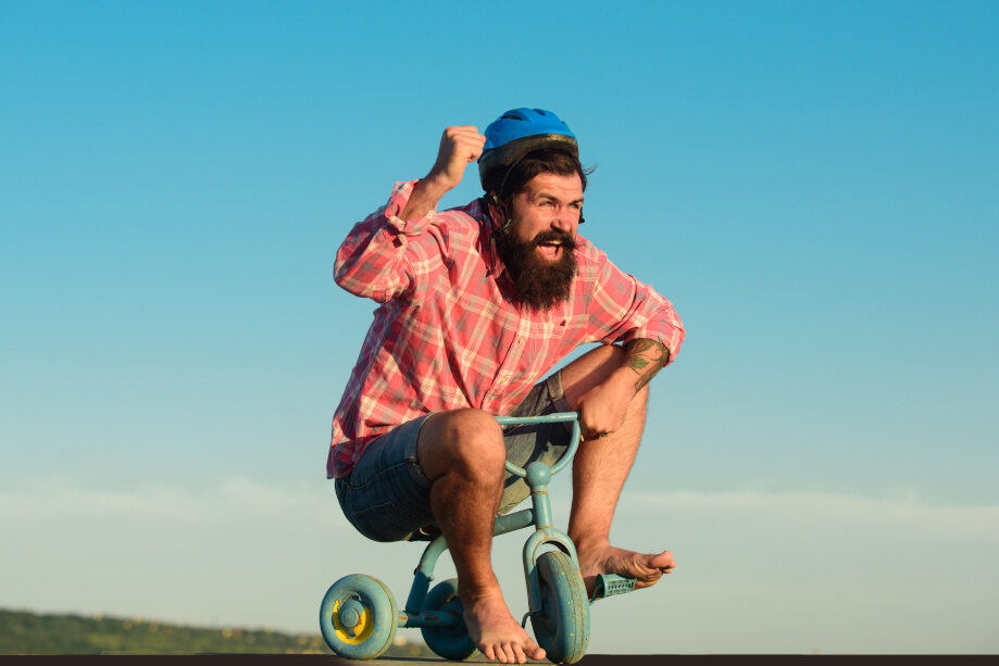 Bärtiger Mann fährt lachend und barfuß ein viel zu kleines Kinderfahrrad, trägt dabei einen Helm.