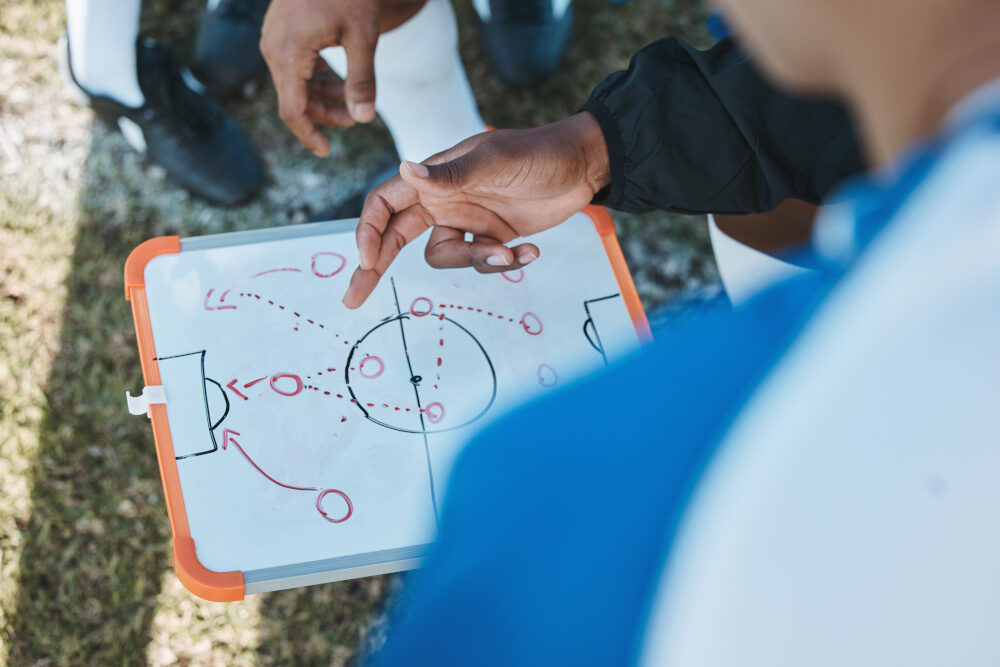 Nahaufnahme einer Taktiktafel mit Skizze eines Fußballfeldes. Hände von Spielern oder Trainern zeigen auf Spielzüge.