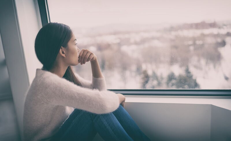 Junge Frau sitzt nachdenklich am Fenster und blickt hinaus auf eine winterliche Landschaft.