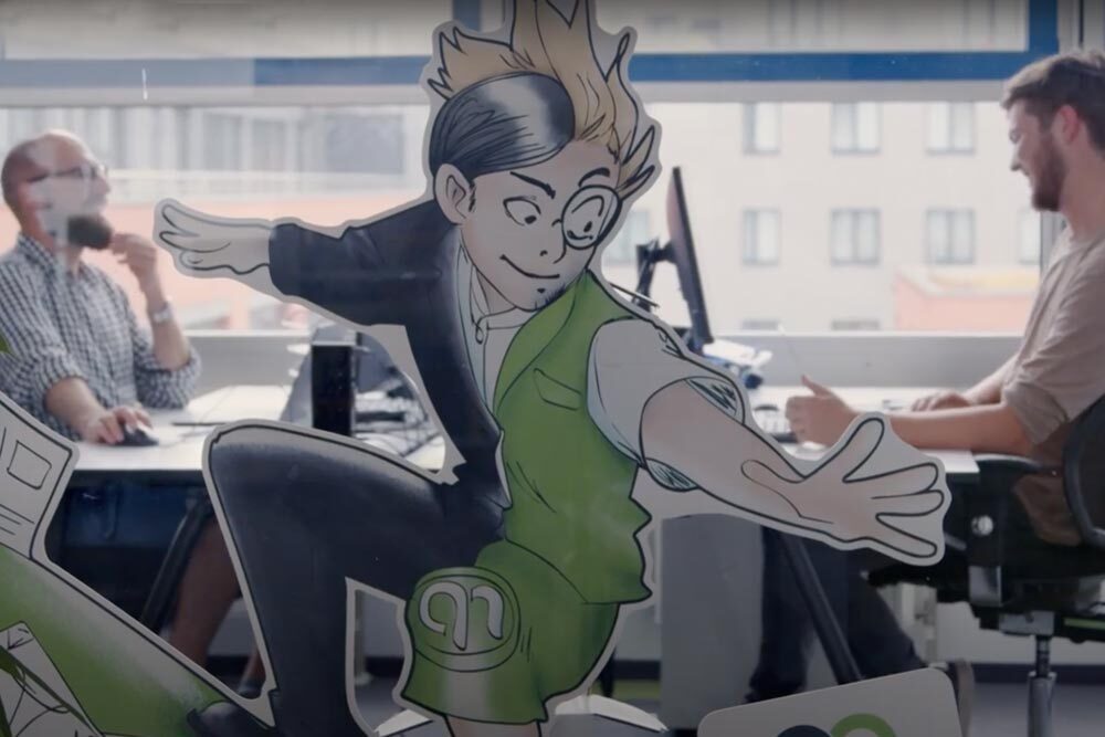 Innenaufnahme eines modernen Büros mit zwei Männern an Schreibtischen im Hintergrund, im Vordergrund eine große bunte Comicfigur auf Glas, die dynamisch nach vorne springt.
