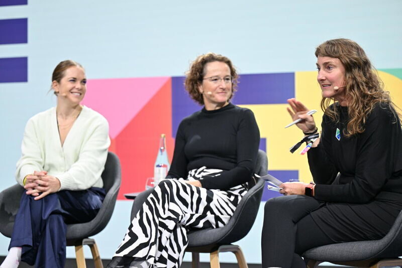 Drei Frauen sitzen auf Sesseln auf einer Bühne vor buntem Hintergrund. Ganz rechts spricht eine Frau mit Mikrofon, sie heißt Insa. Die Frau in der Mitte trägt eine schwarz-weiße Zebra-Hose, die linke Frau trägt einen weißen Pullover. Alle drei wirken engagiert und aufmerksam.
