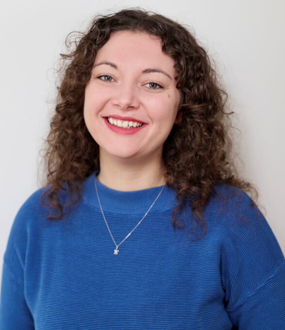 Das Portrait einer lächelnden Frau mit braunen, lockigen Haaren, die einen blauen Pullover und eine silberne Kette trägt