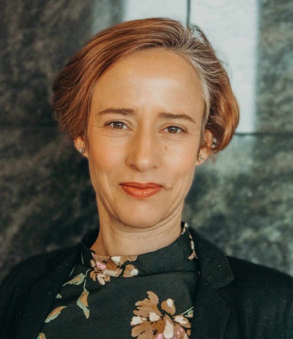 Das Portrait einer Frau mit hellbraunen Haaren, die einen schwarzen Blazer und ein Hemd mit Blumenmuster trägt