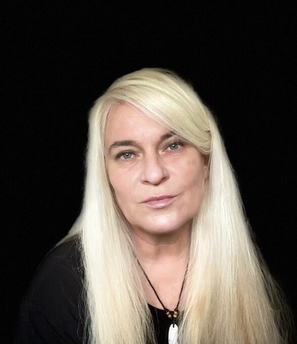 Das Portrait einer Frau mit langen, blonden Haaren. Sie trägt ein schwarzes Oberteil und eine dunkle, dünne Halskette mit einem weißen Anhänger.