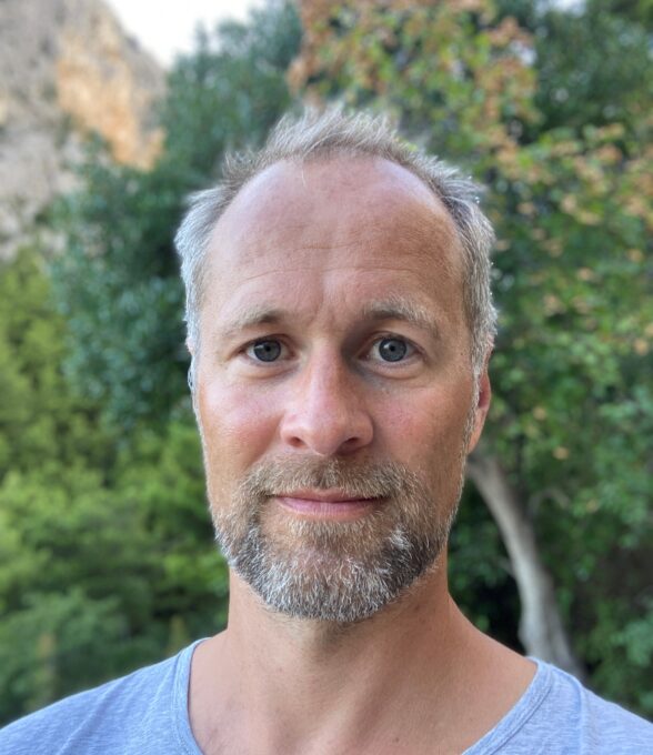 Das Portrait eines Mannes mit grauen Haaren in einem grauen Bart, der ein hellblaues Shirt trägt. Im Hintergrund sind verschwommen Bäume zu sehen.