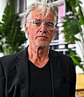 Das Portrait eines Mannes mit grauen Haaren und Brille, der einen schwarzen Blazer mit einem schwarzen Hemd trägt. Im Hintergrund sind verschwommen Zimmerpflanzen in einem Büro zu sehen.