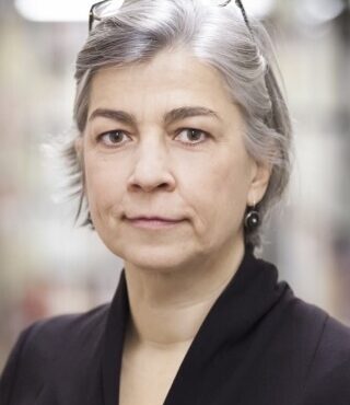 Das Portrait einer Frau mit grauen Haaren. Sie trägt eine Brille auf dem Kopf und einen violetgrauen Blazer.