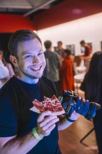 Ein junger Mann lächelt in die Kamera, hält ein Stück Pizza in der einen Hand und eine große Kamera in der anderen. Im Hintergrund sind weitere Personen bei einer Veranstaltung zu sehen.