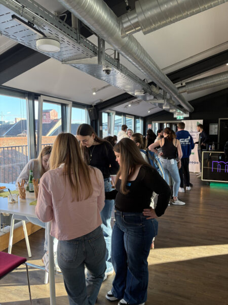 Eine Gruppe junger Menschen steht und unterhält sich an einem hohen Tisch in einem hellen Raum mit großen Fenstern. Auf dem Tisch stehen Getränke und Snacks, im Hintergrund weitere Teilnehmende.
