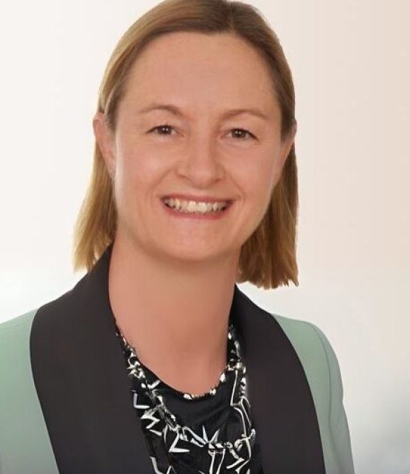 Frau mit kurzem blondem Haar, hellem Blazer und gemustertem Oberteil, lächelt in die Kamera.