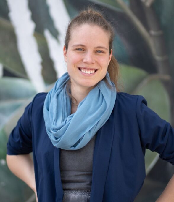 Frau mit Pferdeschwanz, blauem Schal und dunkelblauer Jacke, steht vor einem Wandbild.