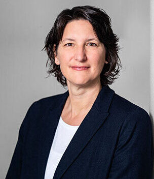 Frau mit dunklen kurzen Haaren, dunklem Blazer und weißem Shirt vor neutralem Hintergrund.