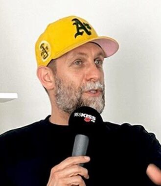 Mann mit Bart und gelbem Cap spricht in ein Mikrofon, neutrale Wand im Hintergrund.