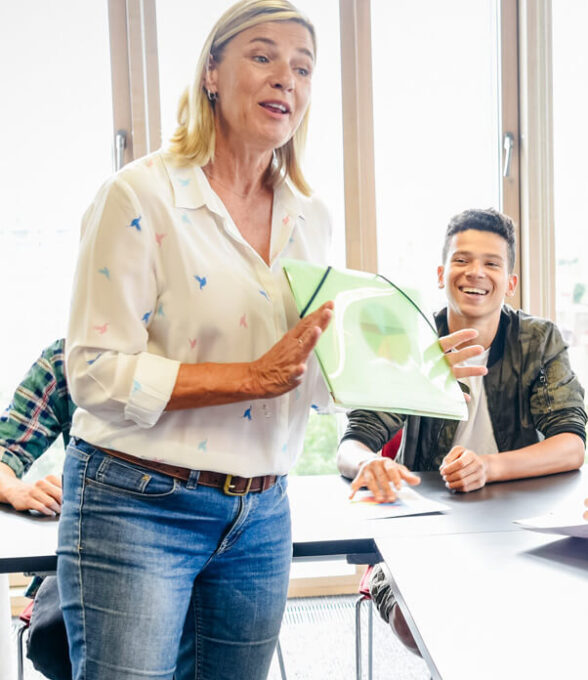 Eine Lehrerin steht vor der Klasse, hält eine Mappe hoch und spricht zu den Schülern, die aufmerksam zuhören.