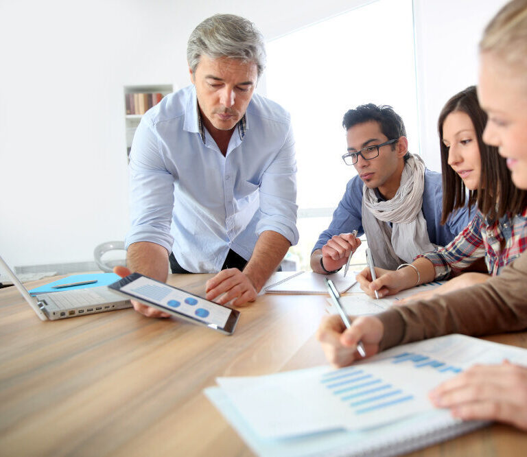 Ein Medienprofi erklärt drei Schüler:innen mit Hilfe eines Tablets und Diagrammen auf Papier wirtschaftliche Zusammenhänge in einer Lernsituation.