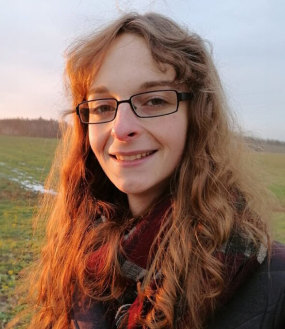 Alana Friedrichs steht im Freien mit langen, lockigen, braunen Haaren. Sie trägt eine Brille und einen Schal. Im Hintergrund ist eine grüne Wiese und ein bewölkter Himmel zu sehen.