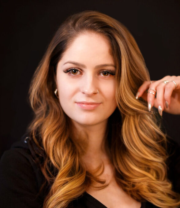 Ann Kathrin Metzler mit langen, welligen, kastanienbraunen Haaren. Sie trägt ein schwarzes Oberteil und hat eine Hand mit langen Nägeln an ihrem Kinn.