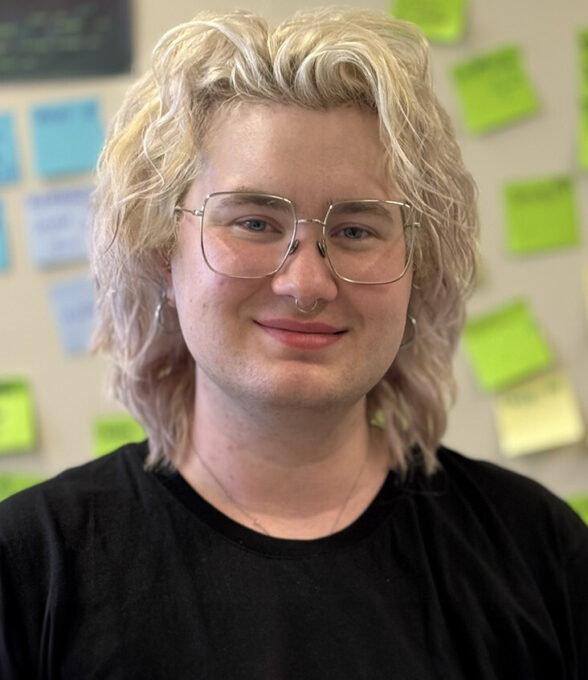 Beck Niederlaender mit blonden, lockigen Haaren, trägt ein schwarzes T-Shirt. Im Hintergrund sind bunte Haftnotizen an einer Wand angebracht.