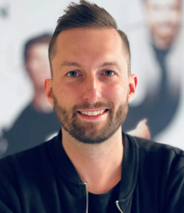 Dominik Koislmeyer trägt einen schwarzen Reißverschluss-Pullover und sitzt vor einem verschwommenen Hintergrund mit weiteren Personen.