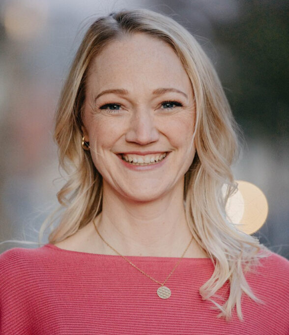 Eva Walig trägt ein rotes Oberteil und eine goldene Halskette. Sie hat lange, blonde Haare und steht im Freien mit unscharfem Hintergrund.