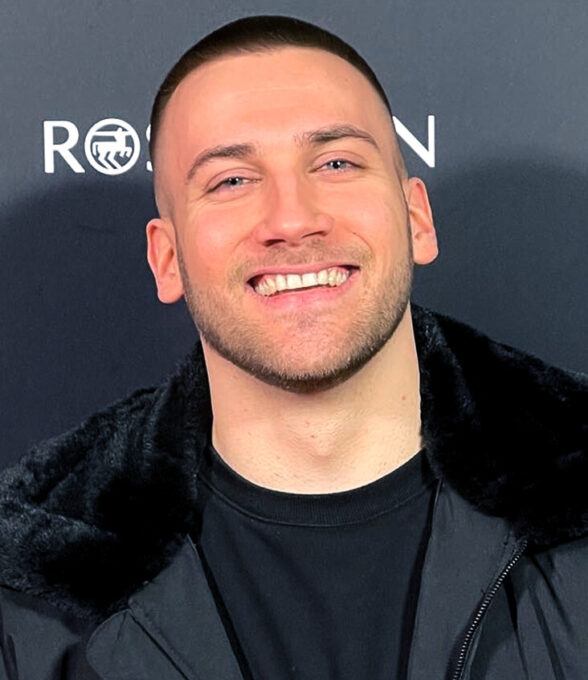 Floder Flo mit kurzem Haar trägt ein schwarzes Oberteil und eine schwarze Jacke mit Pelzkragen. Hintergrund mit dem Logo von 'ROTTEN'.