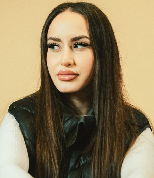 Joyyy, eine Comedien TikTokerin, trägt eine schwarze Weste und hat lange, glatte Haare. Sie sitzt mit verschränkten Armen.