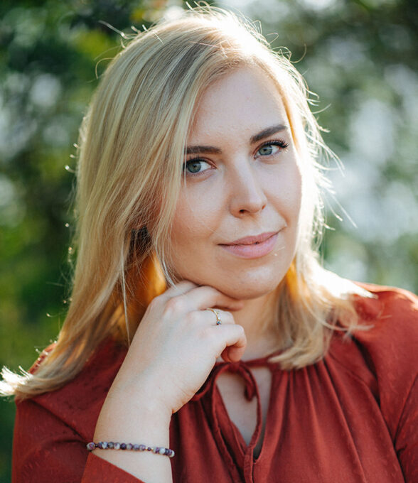 Katarzyna Karpinska mit blonden Haaren, trägt ein rotes Oberteil, sitzt im Freien und hält ihre Hand an das Kinn.