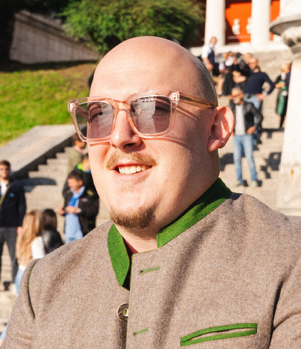 Mann mit Glatze und Brille trägt eine graue Jacke mit grünen Akzenten. Im Hintergrund sind Menschen auf einer Treppe zu sehen.