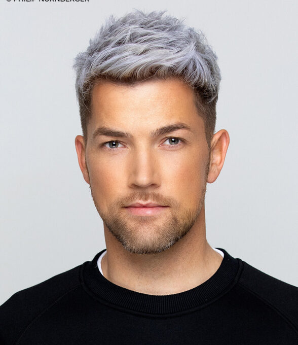 Auf dem Bild ist ein professionelles Porträt von Sebastian Schilde, einem Musikproduzenten, zu sehen. Er trägt ein schwarzes T-Shirt, hat graue Haare und einen Bart.