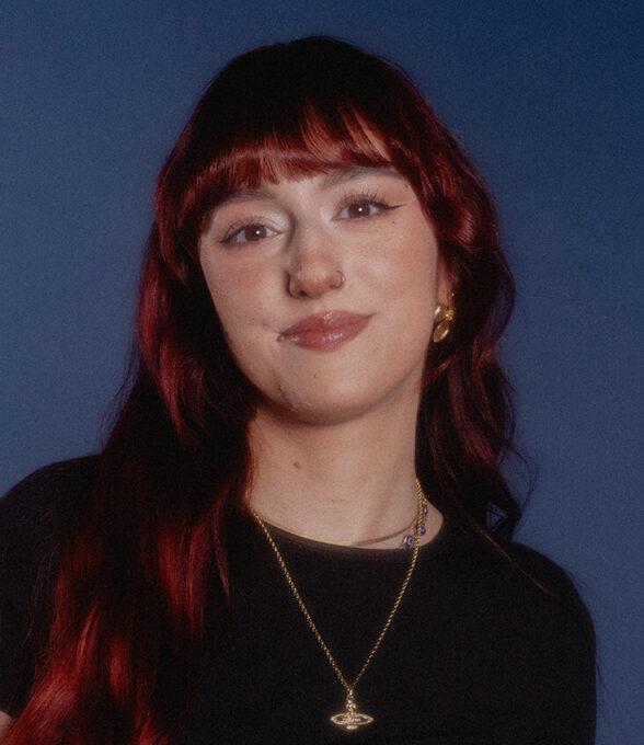 Sejla Begovic trägt ein schwarzes Oberteil und eine goldene Halskette mit einem Saturn-Anhänger. Ihre langen, rotbraunen Haare fallen über die Schultern.