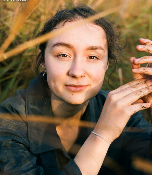 Swantje Furtak sitzt inmitten hoher, goldener Gräser und blickt ruhig und direkt in die Kamera. Ihre Hände, leicht mit Erde verschmiert, hält sie entspannt vor sich; ihr Gesicht wird von warmem Sonnenlicht beleuchtet. Die natürliche Umgebung und das sanfte Licht verleihen dem Bild eine ruhige, erdverbundene Stimmung.