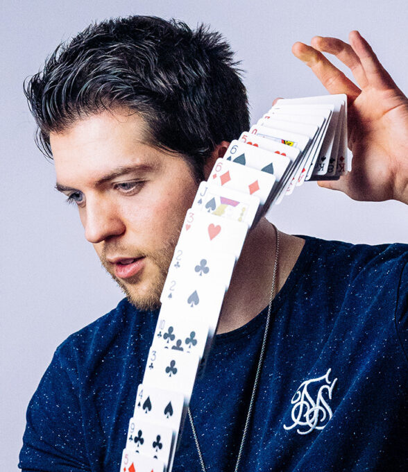 Alexander Straub, ein Magier, hält eine fächerförmig angeordnete Karte in der Hand. Er trägt ein dunkelblaues T-Shirt mit einem weißen Logo.