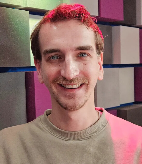 Andre Wengenroth lächelt freundlich in die Kamera. Er trägt ein helles Sweatshirt und steht vor einer Wand mit farblich abgesetzten, schallabsorbierenden Paneelen. Sanftes rotes Licht fällt auf seine Haare und Schultern.