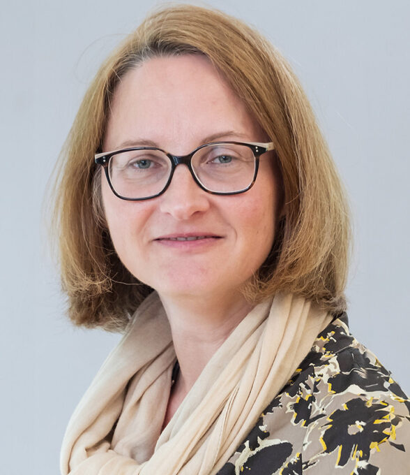 Anette Kolb mit schulterlangen, blonden Haaren, trägt einen hellen Schal und eine Bluse mit floralem Muster. Sie schaut leicht zur Seite.