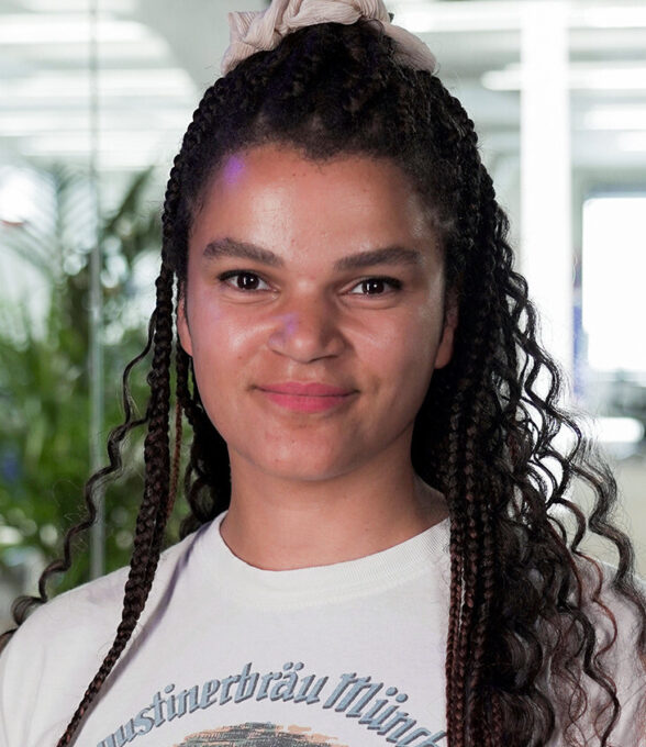 Beatrice Osuji 2 trägt ein weißes T-Shirt mit dem Aufdruck 'Augustiner Bräu München'. Ihre langen, lockigen Haare sind zu Zöpfen geflochten.