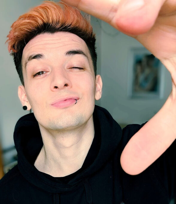 Künstler, Autorund Content Creato Darkvictory mit kurzem, orangefarbenem Haar trägt einen schwarzen Hoodie. Er zeigt mit der Hand auf die Kamera. Hintergrund unscharf.